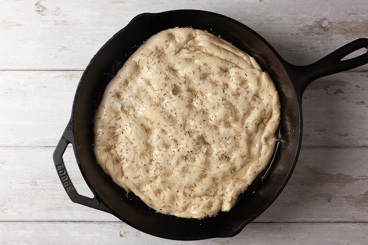 Overhead shot of a cast iron skillet filled with uncooked focaccia dough topped with herbs, resting on a light wooden surface;