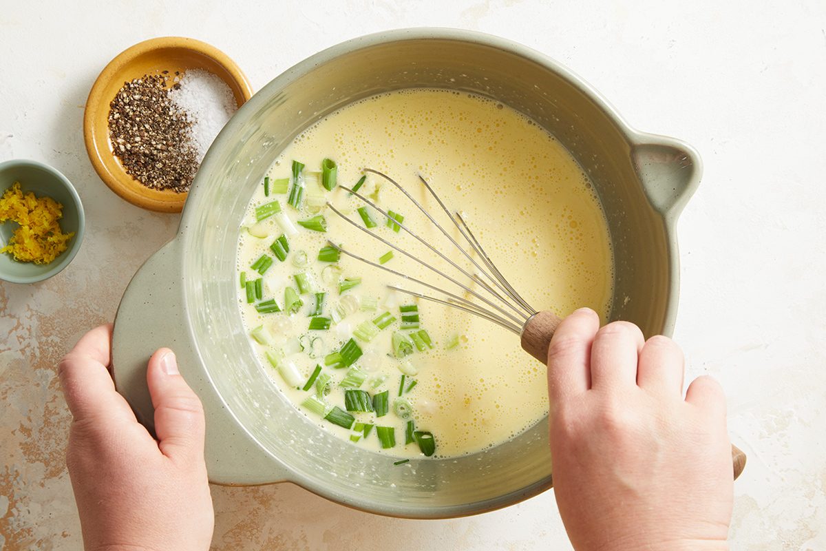 A person uses a whisk to mix chopped green onions into a creamy yellow batter in a bowl. Nearby, small dishes hold salt, pepper, and lemon zest.