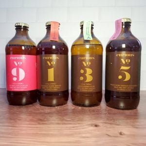 Four brown glass bottles of Curious Elixirs drinks, labeled No. 9, No. 1, No. 3, and No. 5, are lined up on a wooden surface against a white tiled background.