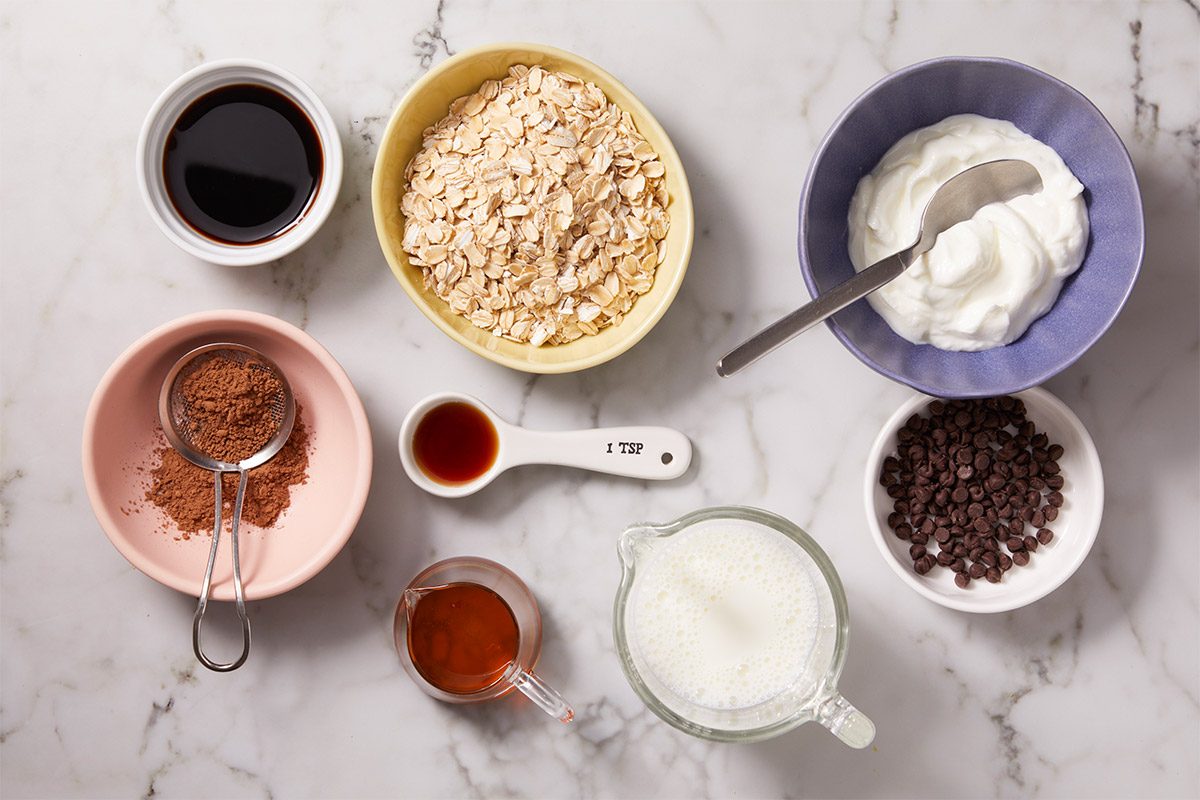 Overhead shot of ingredients arranged on a marble surface, including rolled oats, Greek yogurt, brewed coffee, cocoa powder, maple syrup, vanilla extract, milk, and chocolate chips in small bowls.