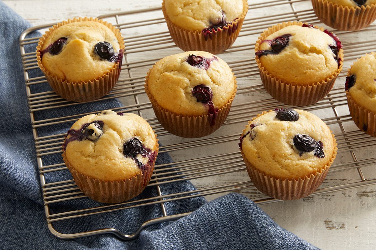 Seven golden-brown blueberry muffins in paper liners cool on a wire rack, with a dark blue cloth underneath, set on a light wooden surface.