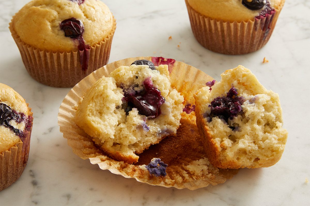 A blueberry muffin split in half on a brown paper wrapper, with whole blueberries visible inside. Two whole muffins are in the background, all on a light-colored, marble-like surface.