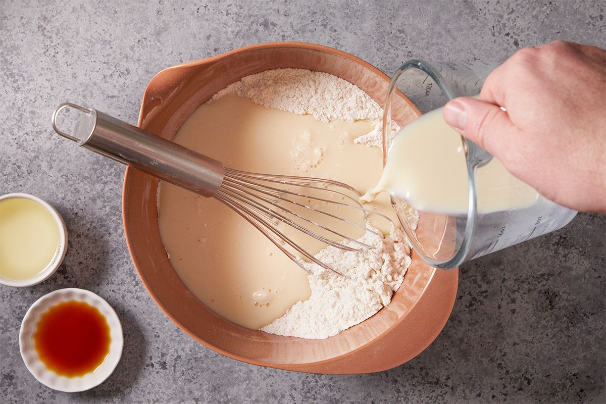 Overhead shot of Milk flows from a glass measuring cup into a mixing bowl with flour and other wet ingredients A whisk sits inside the bowl nearby small bowls of oil and vanilla rest on a gray countertop