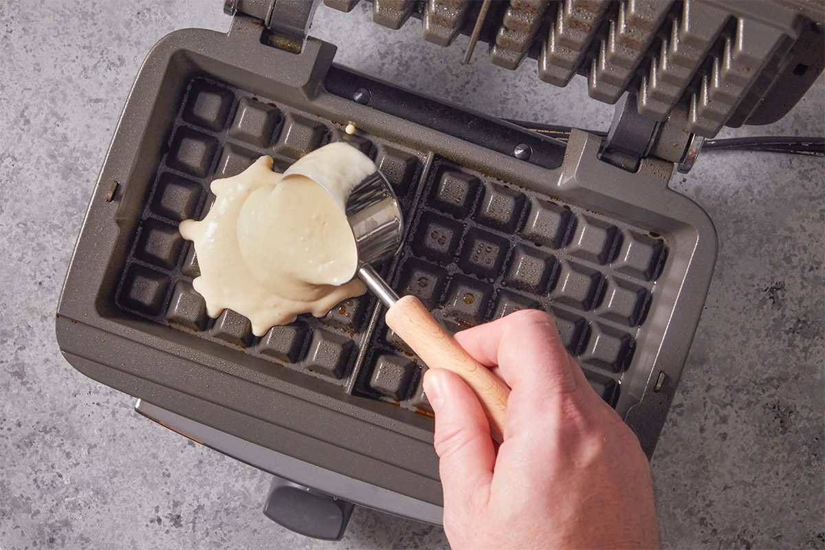 Overhead shot of a hand pours waffle batter from a scoop onto a hot waffle iron The nonstick grid awaits as waffles are about to be made
