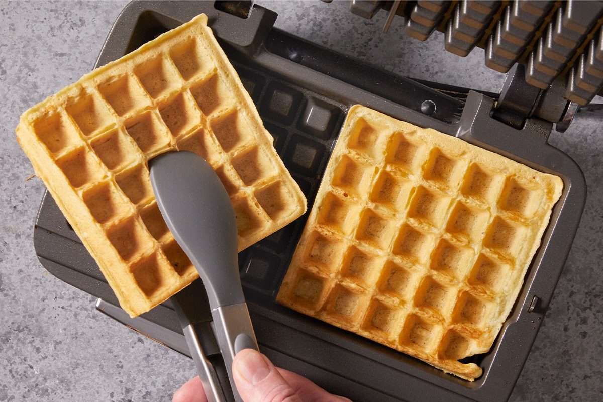 Overhead shot of a hand lifts a golden brown waffle from a waffle maker using tongs as another waffle cooks inside the appliance on a gray countertop