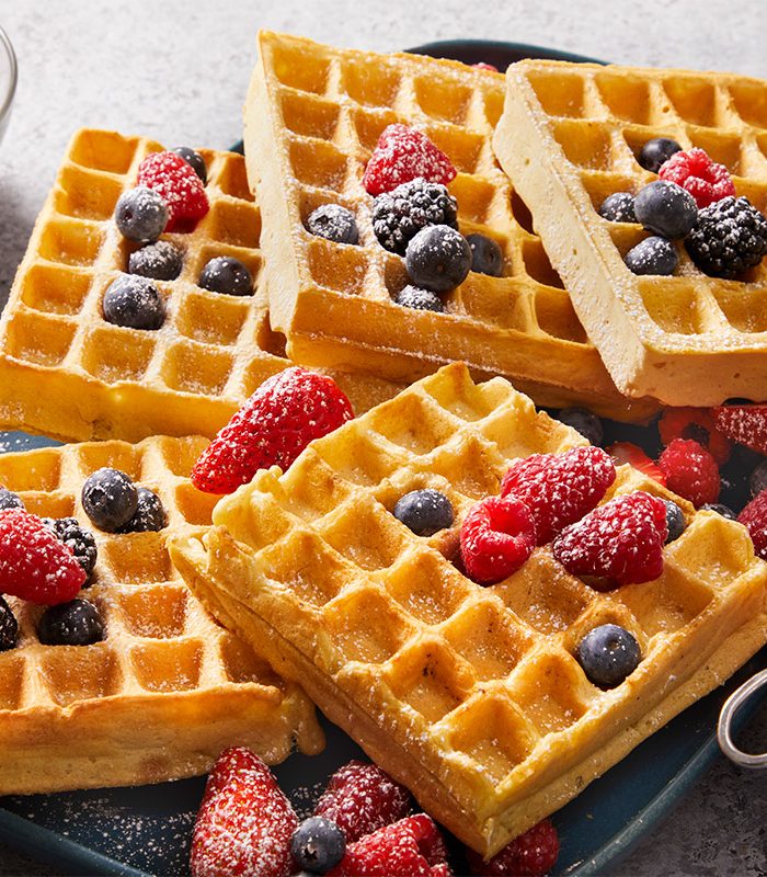3/4th Shot of Vegan Waffles are served on a plate with fresh blueberries raspberries and strawberries gently dusted with powdered sugar Nearby there is a bowl of mixed berries and a small dish holds a sifter