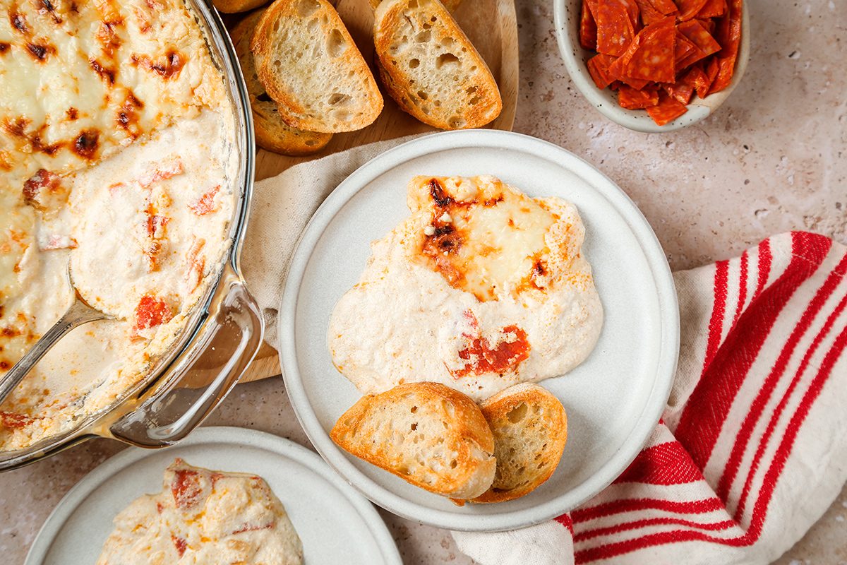 A plate with creamy, cheesy dip topped with tomato, served with slices of toasted baguette. Nearby, there is a dish of dip, a bowl of pepperoni, extra bread slices, and a red-and-white striped towel.